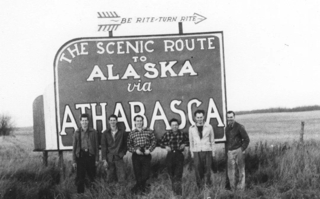 Black and white photograph taken in 1948 of a group of six men standing in front of a large highway sign on the edge of a field that reads: Be Rite – Turn Rite. The Scenic Route to Alaska via Athabasca