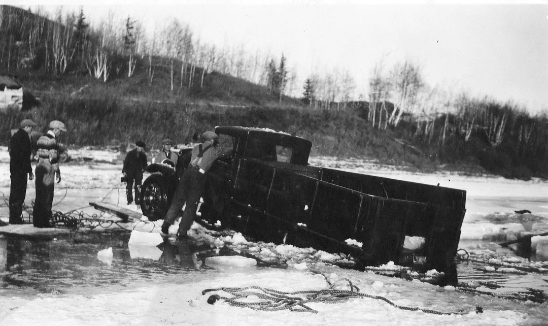 Black and photograph of a 1930s-era panel truck half submerged in an icy river. A man is standing on a block of ice, reaching into the cab. Four other men are standing on the ice, looking on and holding cables and ropes.