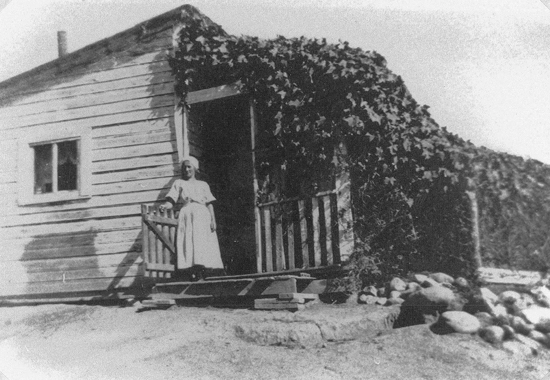 Black and white photograph of a woman standing on the steps of a wood-framed dwelling that is covered by ivy on one side.
