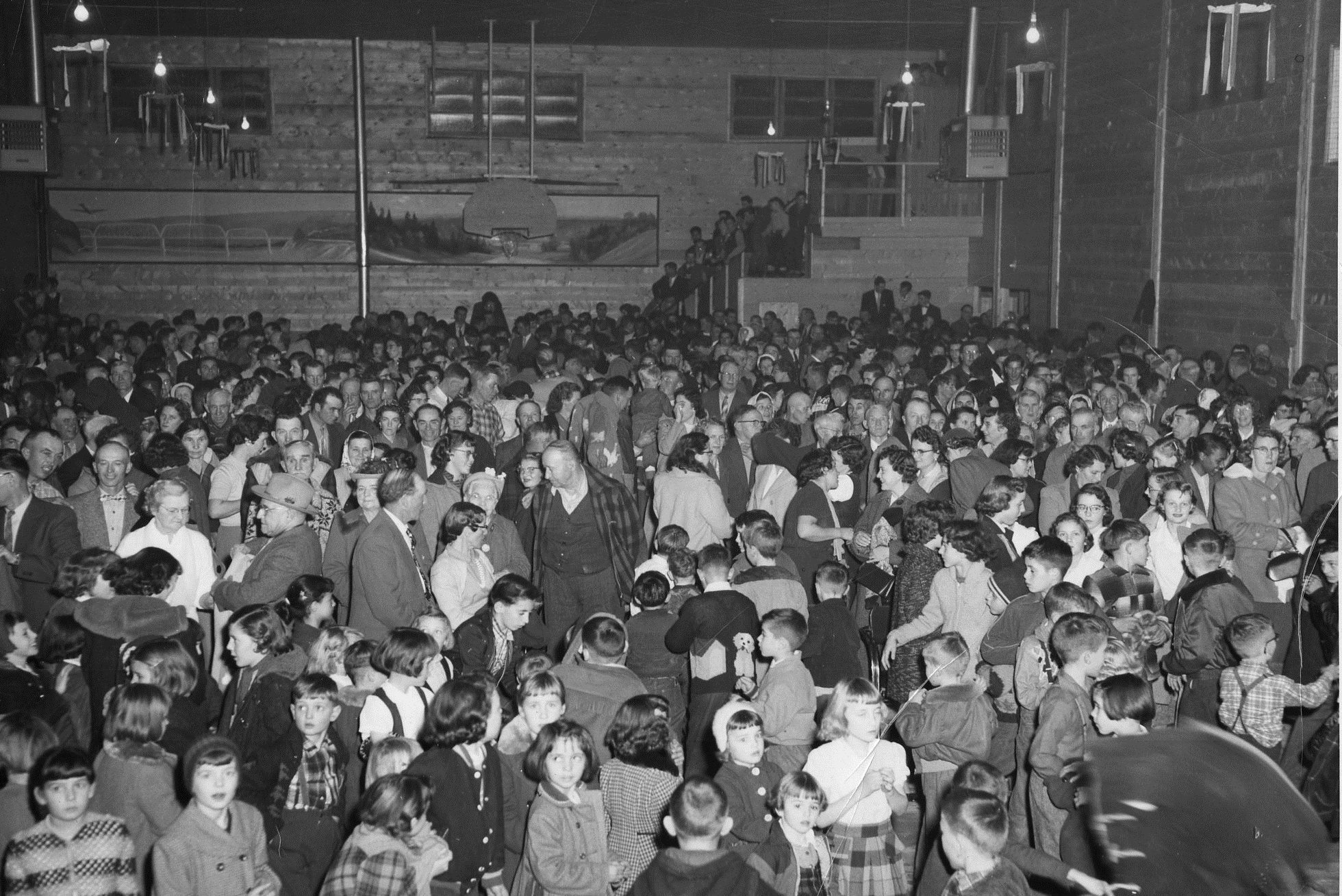 Black and white photo taken inside a large wood-framed building of a very large group of people. In the foreground are fifty or more children dressed in good clothing. 