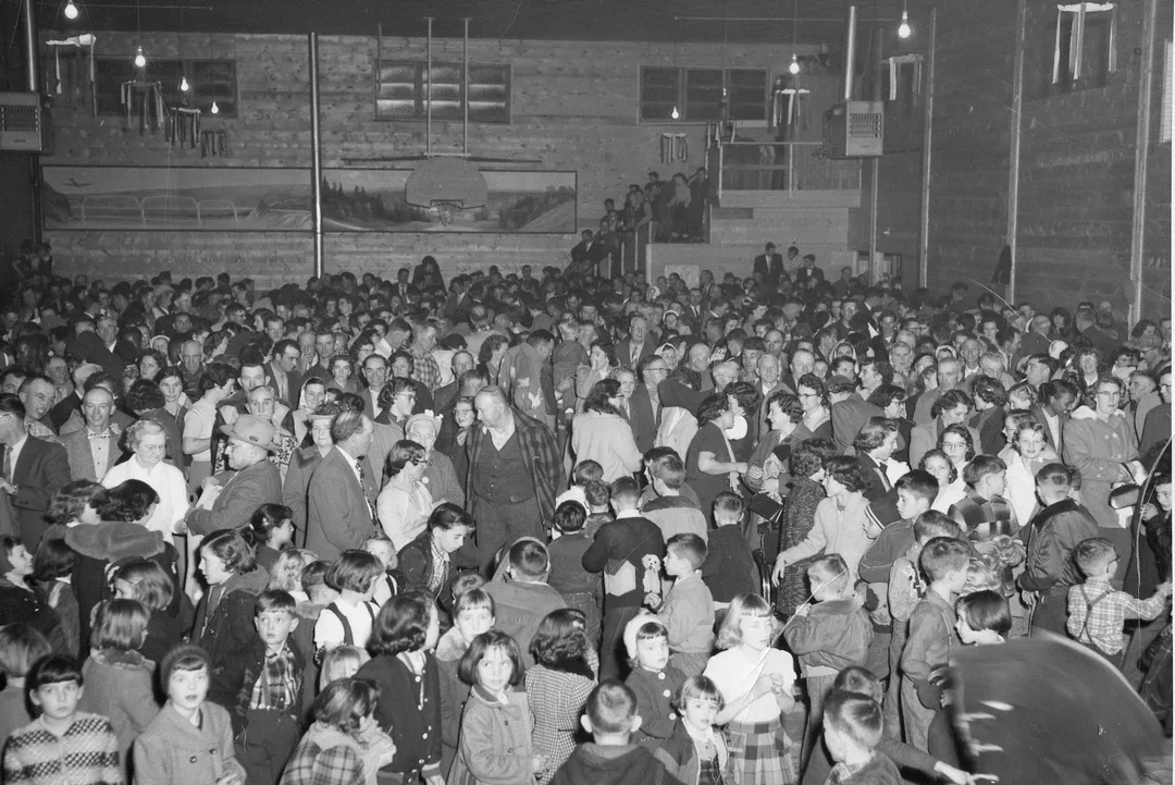 Black and white photo taken inside a large wood-framed building of a very large group of people. In the foreground are fifty or more children dressed in good clothing. 