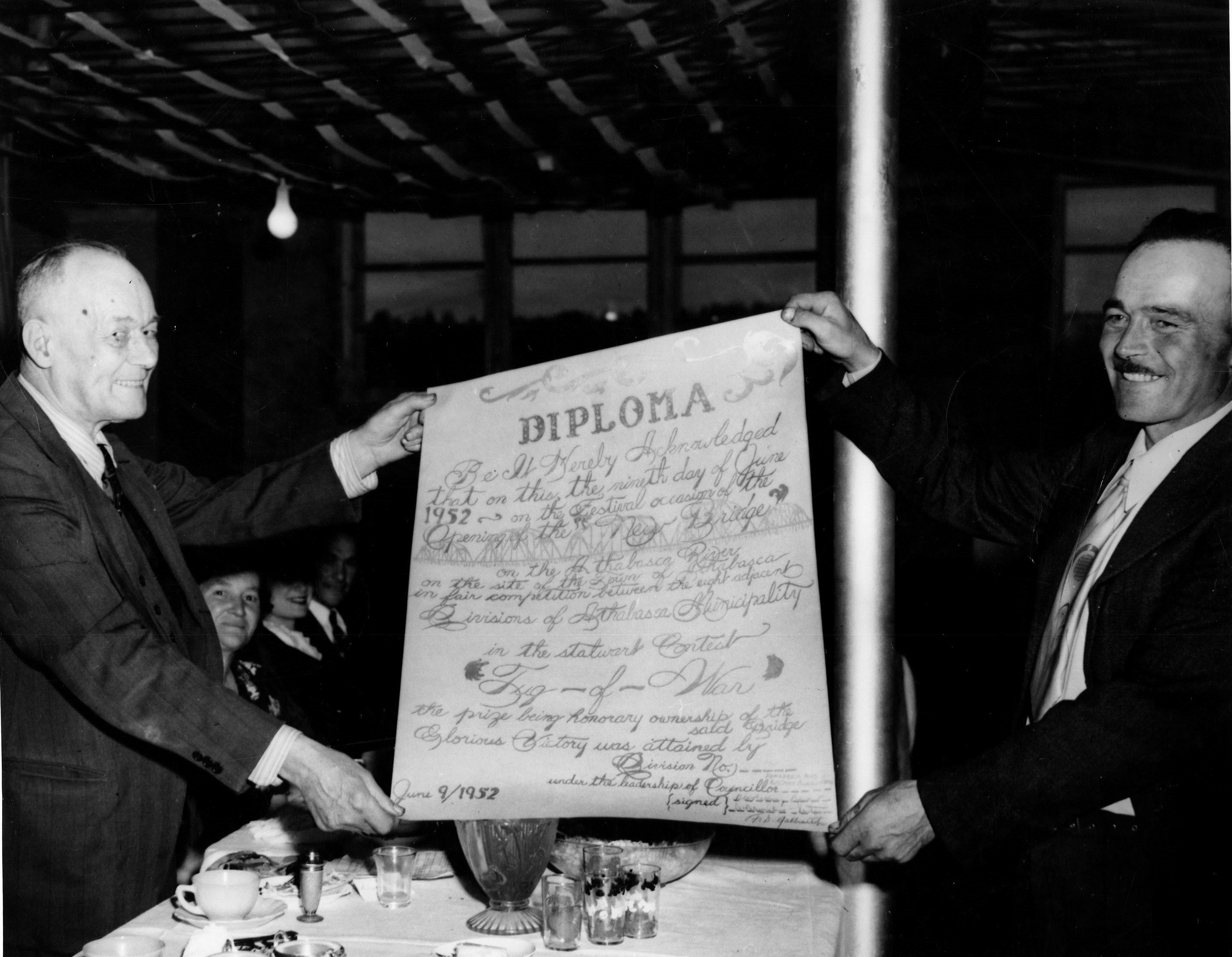 Black and white photo of two men holding up a certificate that is labeled DIPLOMA which reads Be it heareby acknowledged that on this the nineth day of June 1952 on the festival ocassion of the opening of the new bridge on the Athabasca River on the site of the Town of Athabasca, etc.
