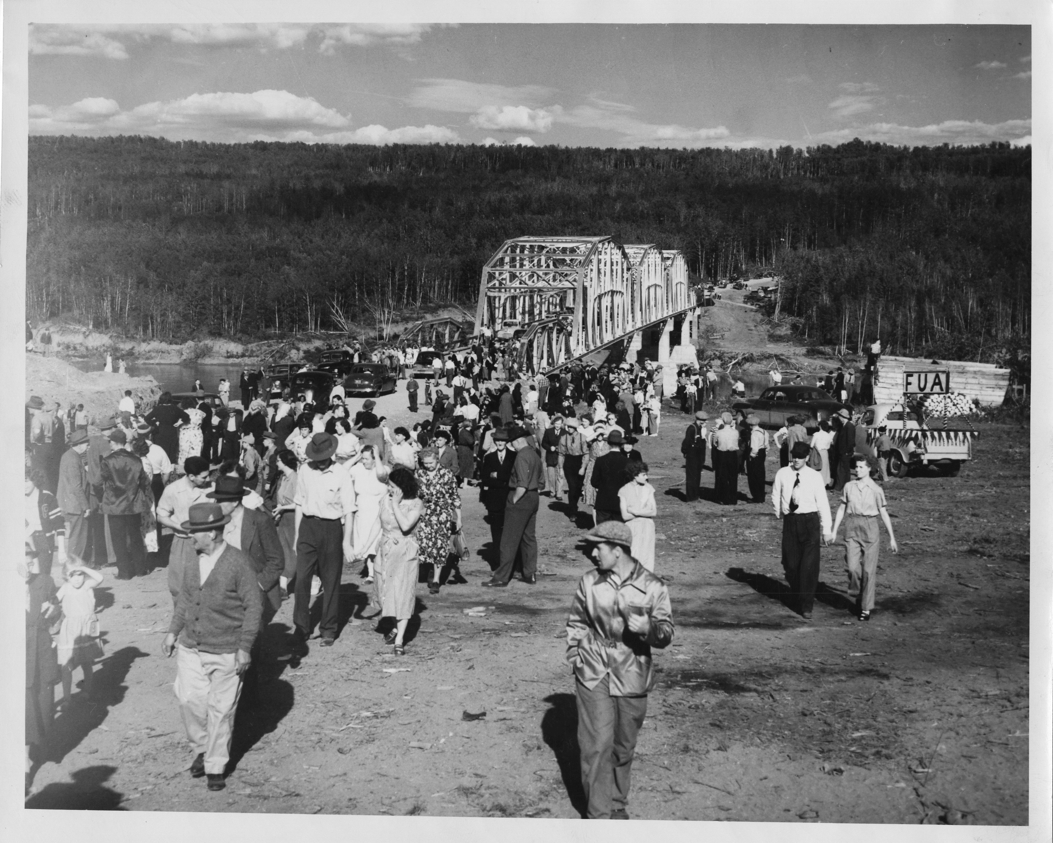 Black and white photograph of a throng of people dresed in light clothing walking away from a steel-truss bridge across a river valley