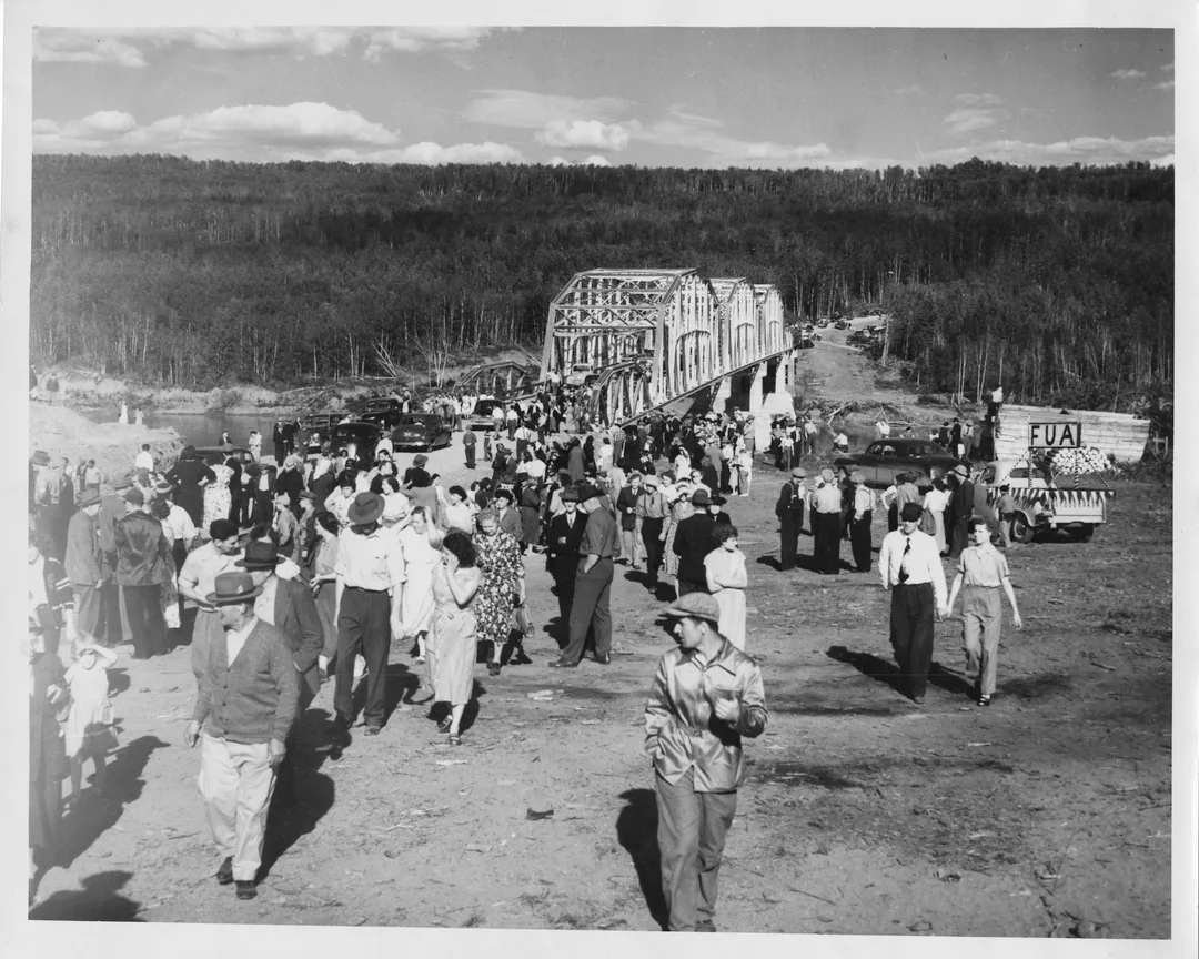 Black and white photograph of a throng of people dresed in light clothing walking away from a steel-truss bridge across a river valley