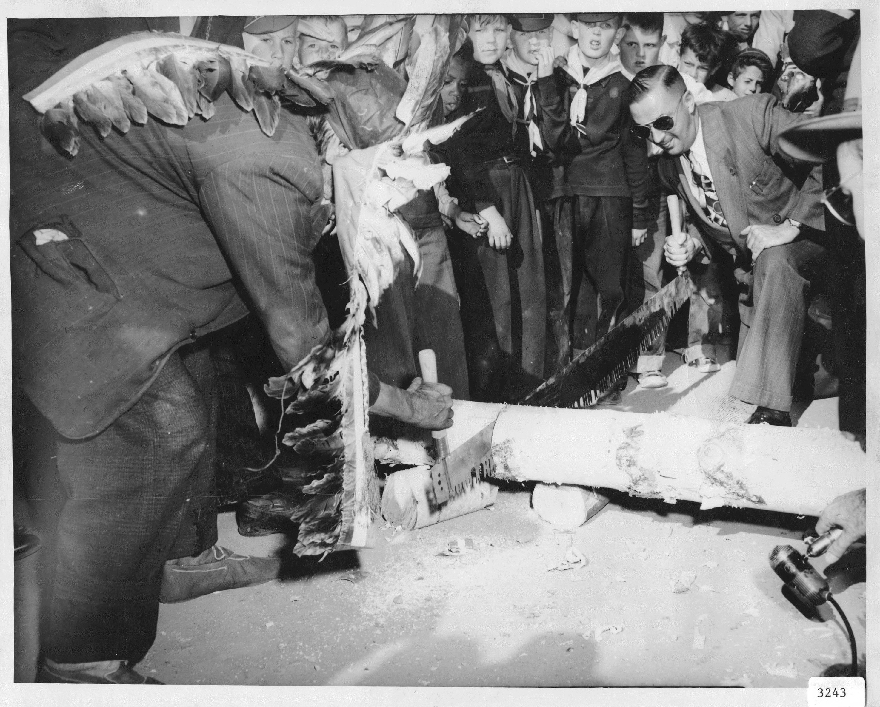 Black and white photo of two men sawing a large log in half. One man is Cree and is wearing a traditional head dress. The other man is Caucasian and is wearing a suit and sunglasses. There is a group of boy scouts in the background looking on.