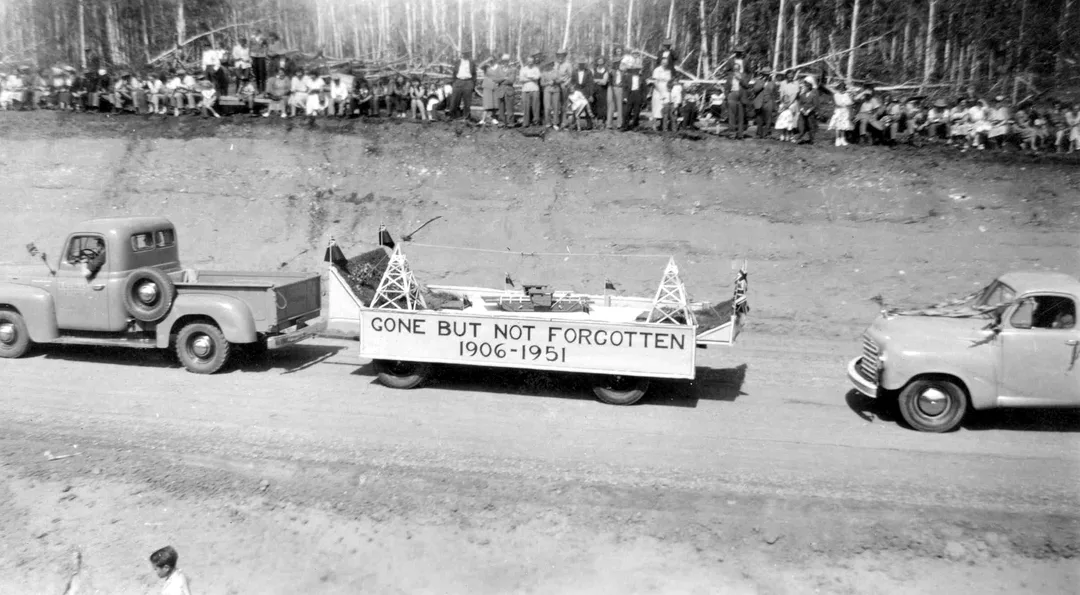 Black and white photo of a 1940s era pick-up truck pulling a flatbed trailer in a parade. The trailer is decorated as a parade float with a banner, 