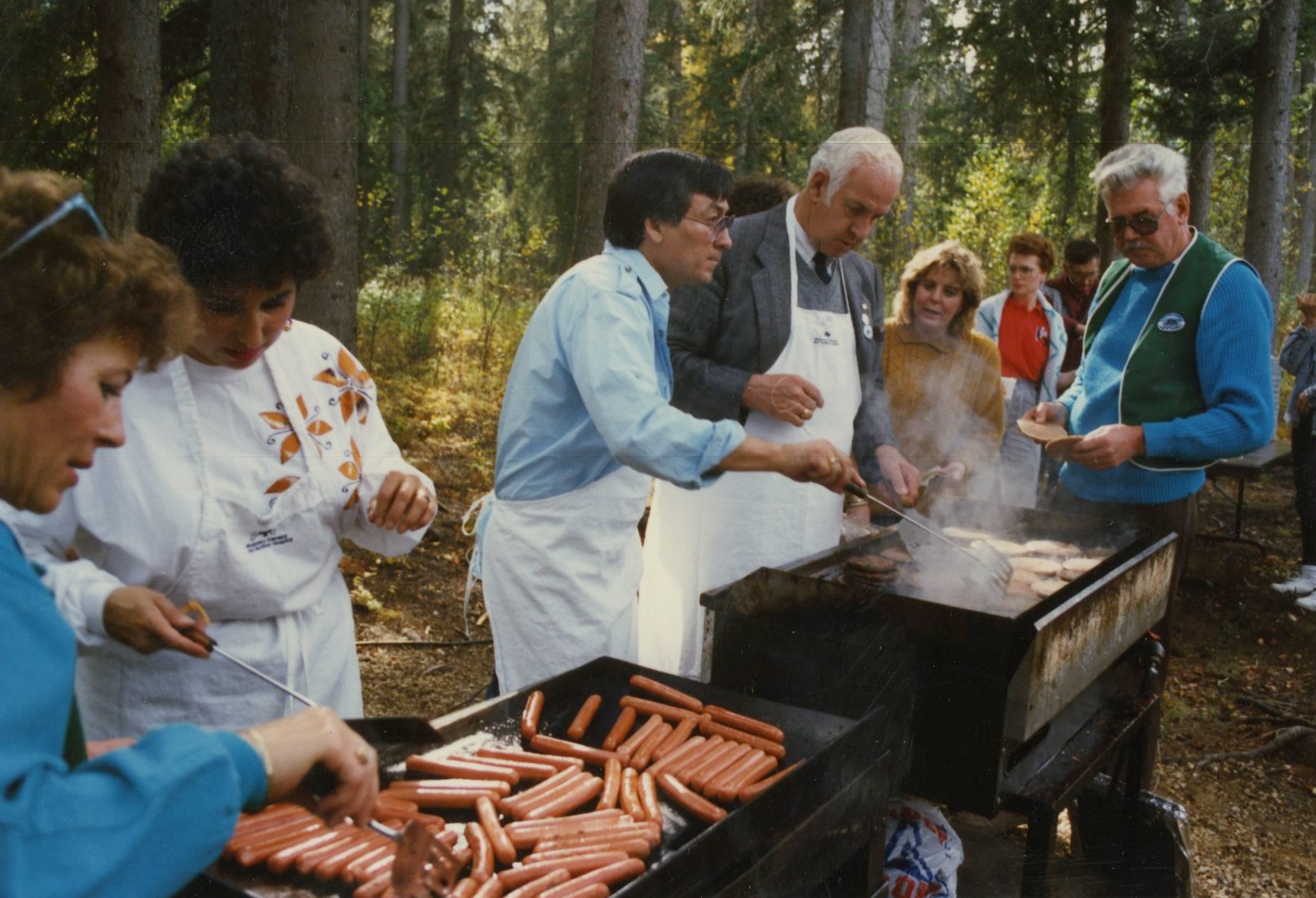 Color photograph of women and men wearing aprons, behind barbeques, cooking hotdogs and hamburgers