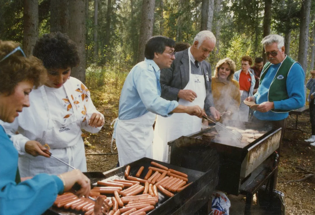 Color photograph of women and men wearing aprons, behind barbeques, cooking hotdogs and hamburgers