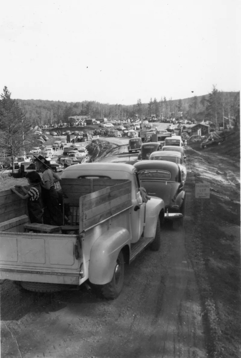 Black and white photo of long lines of 1940s era cars and pick-up trucks parked along a dirt road.