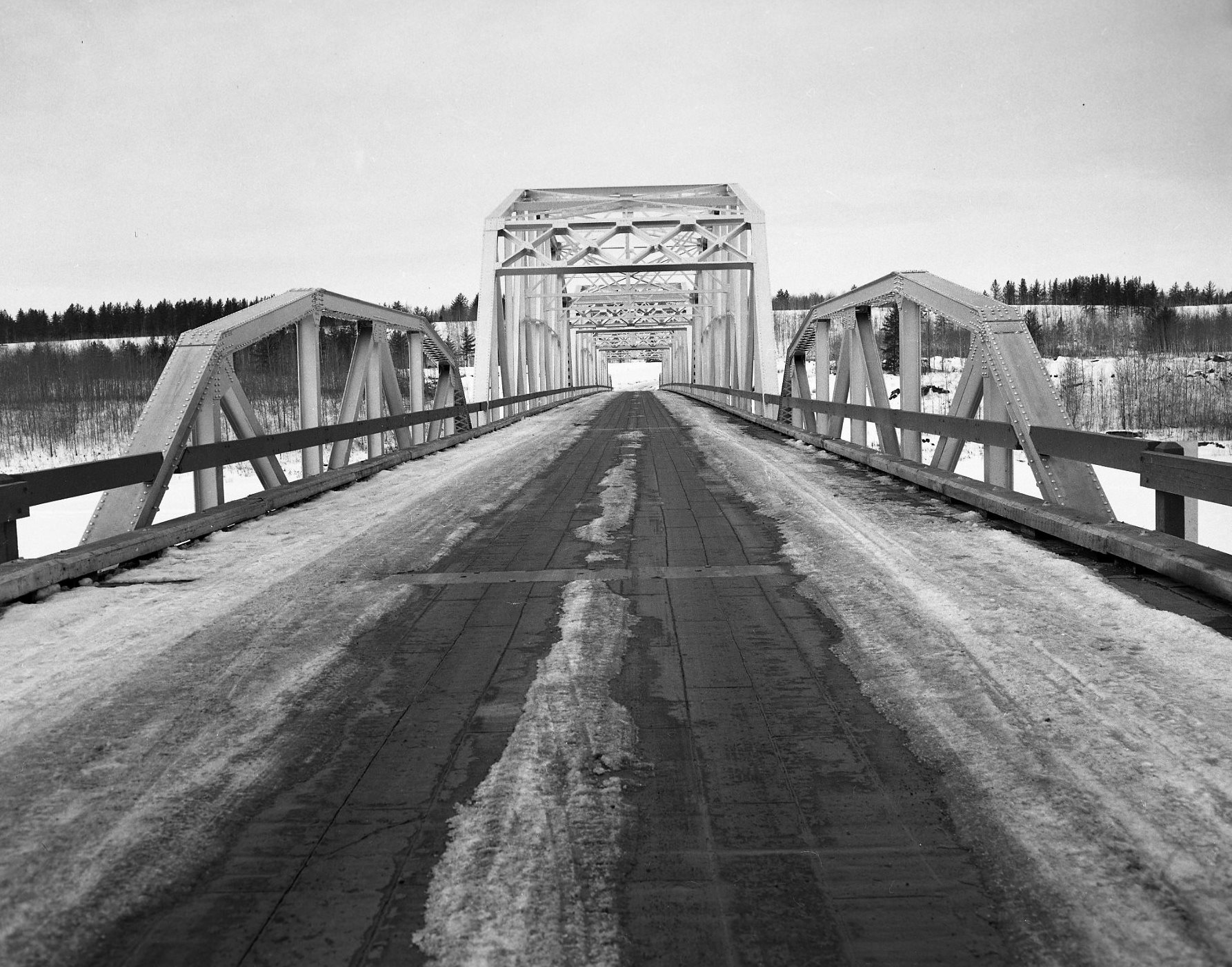 Black and white photograph of a truss-style metal-framed bridge taken from the bridge deck, looking down the length of it