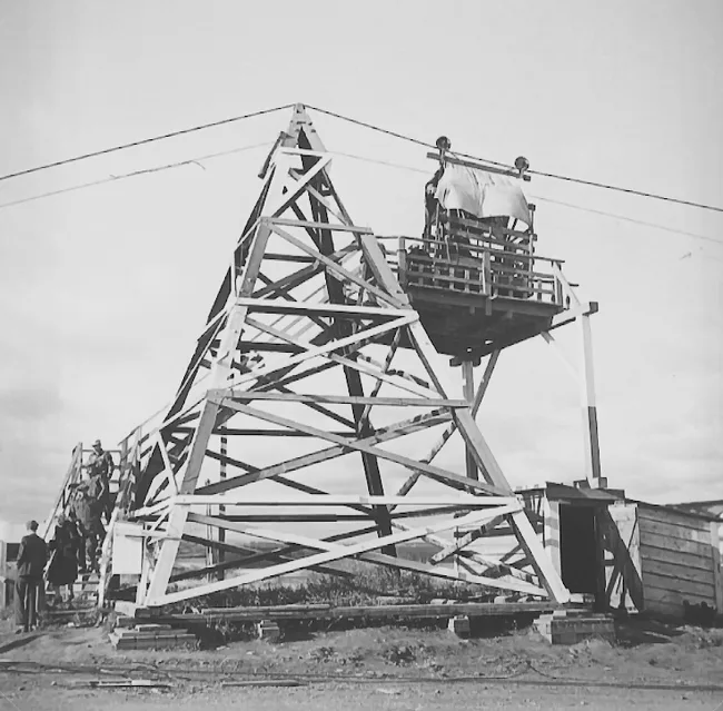Black and white photograph of a wood-framed tower which has a set of stairs wrapping around two sides. People are walking up and down the stairs. There is a platform two thirds of the way up on one side of the tower. From this platform, people are loading into a wood-framed cage that is suspended on two wheels from a pully system, hanging from a cable hung at the top of the tower.